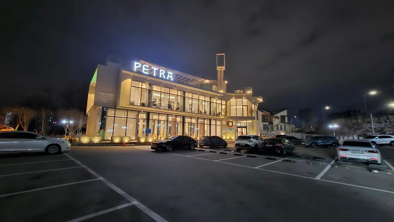 Wurin ajiye motoci na PETRA cafe - faffadan filin ajiye motoci da ginin bene biyu mai gilashi da fitilun dare | 하이제이에스비