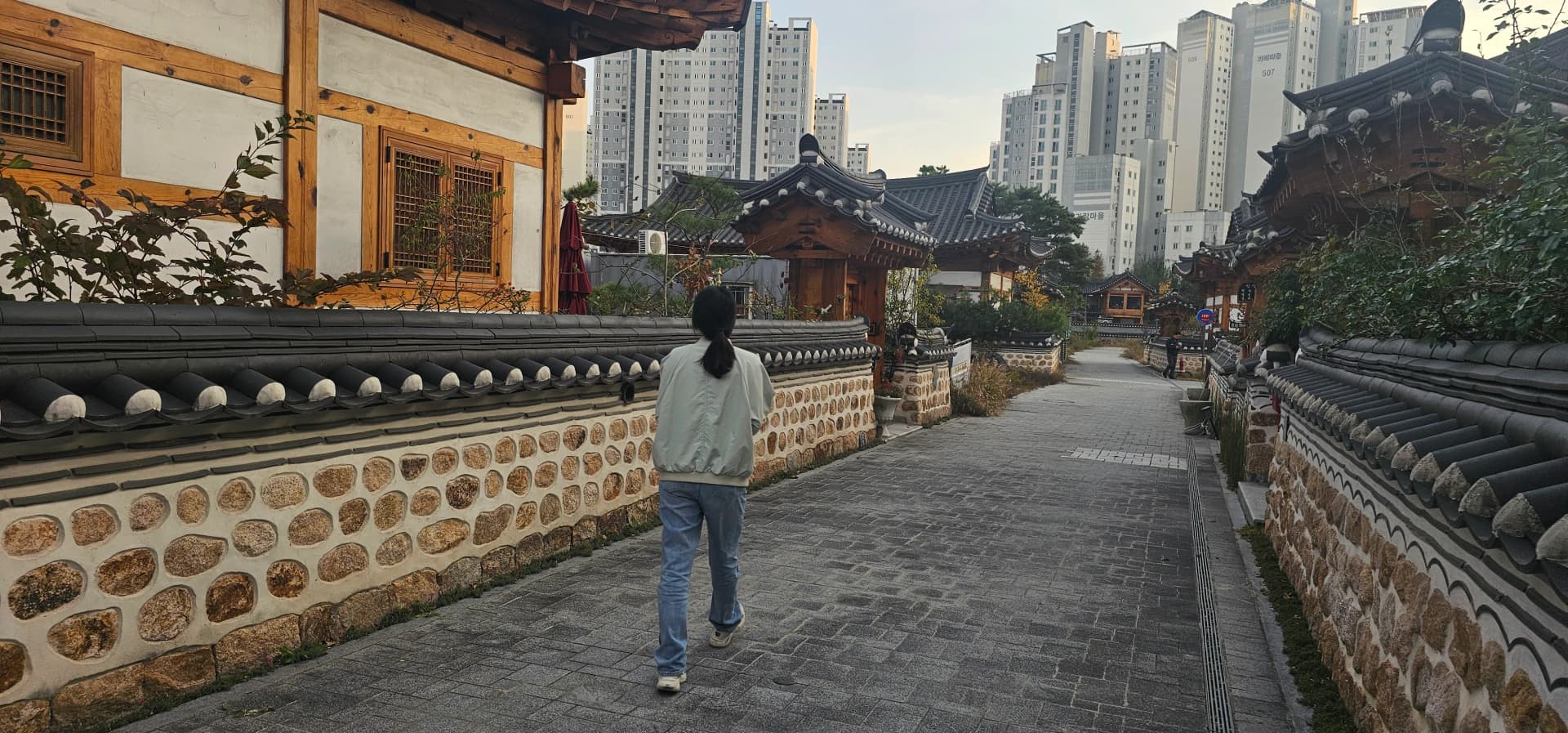 세종 고운동 한옥마을 골목 — 돌담길 양쪽으로 기와지붕 한옥이 늘어서 있고, 뒤로 아파트 단지가 보인다 | 하이제이에스비