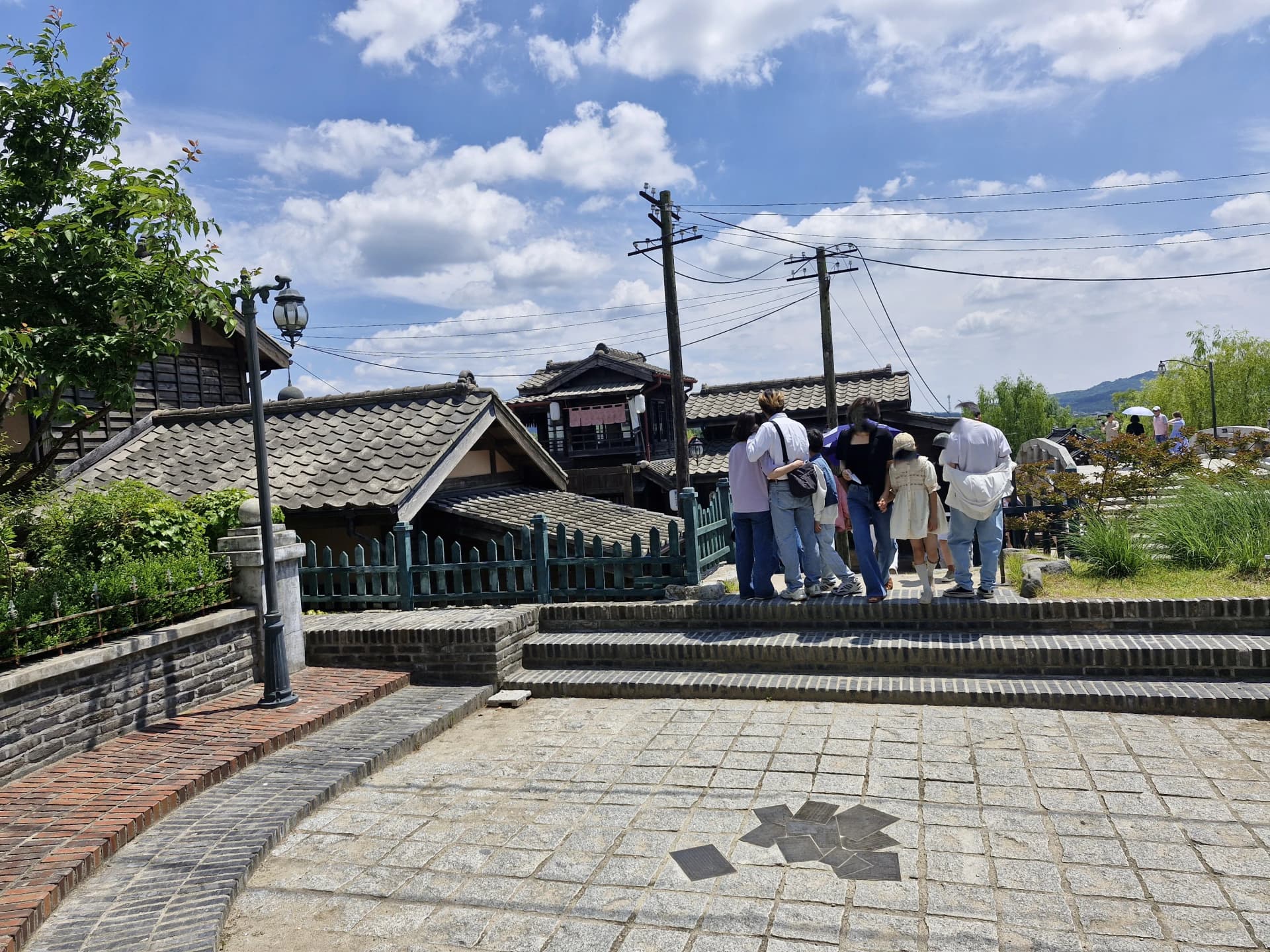 Pengunjung menyeberangi jembatan menuju gang dengan tiang telegraf di latar | 하이제이에스비