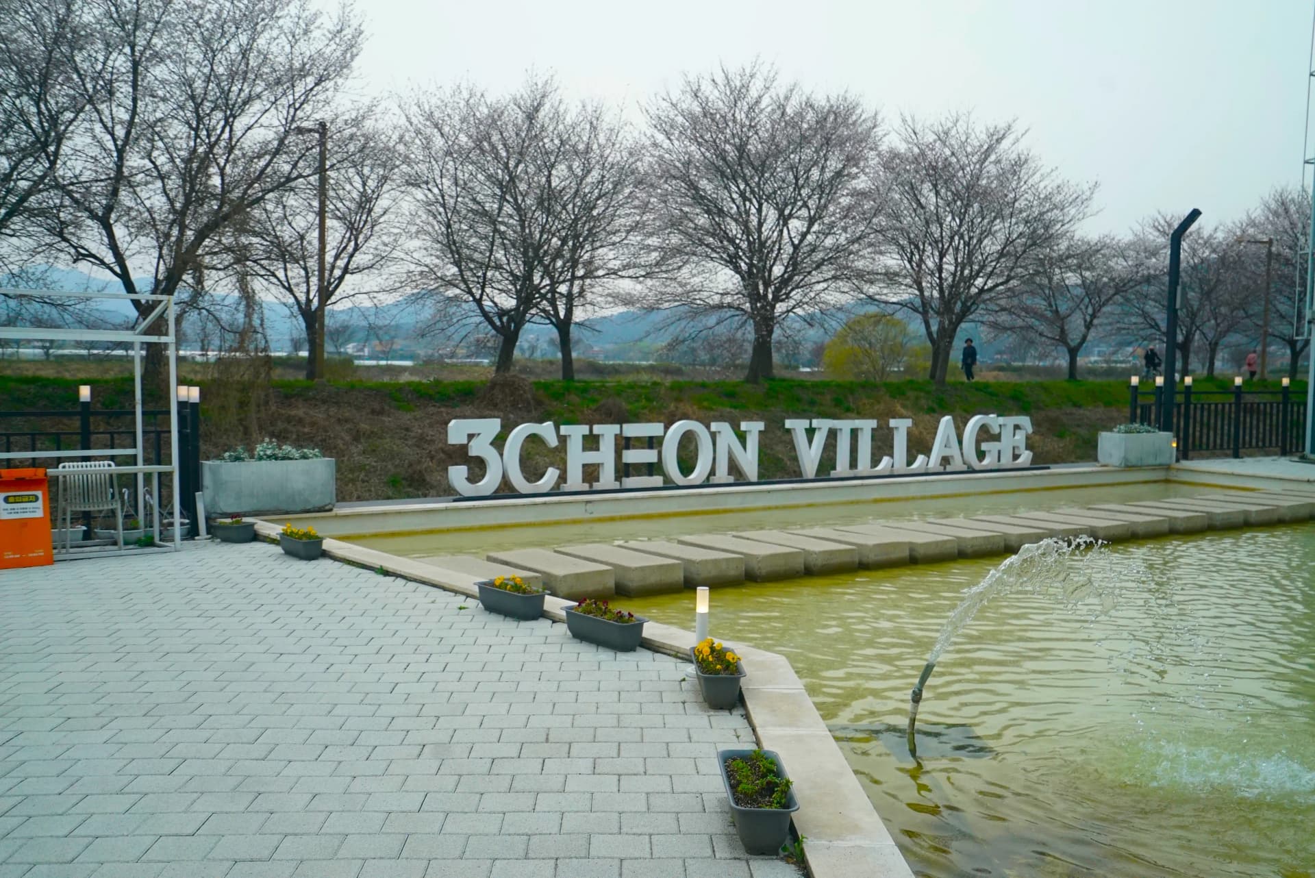 Outdoor plaza with 3CHEON VILLAGE sign and cherry blossom trees lined up in the background