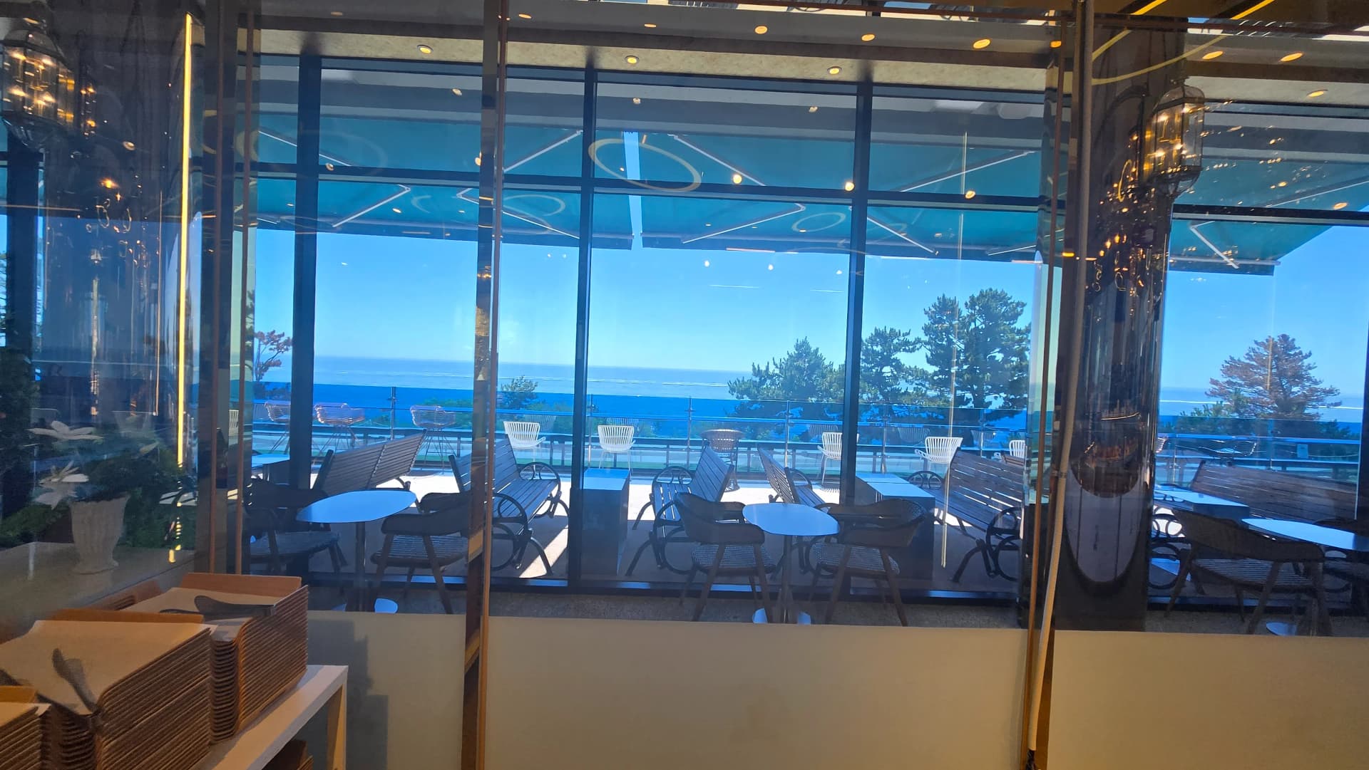 더킹 카페 실내에서 바라본 테라스 좌석과 소나무 너머 동해 수평선 - The King Cafe indoor view of terrace seats with pine trees and East Sea horizon | 하이제이에스비